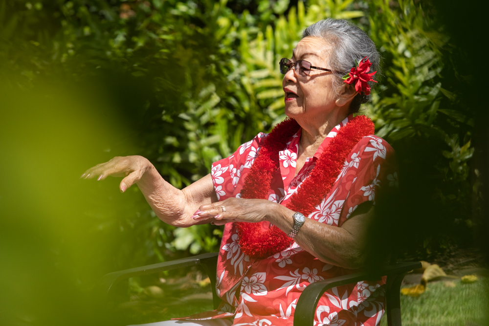 Senior woman performing hula in a garden with a red lei.